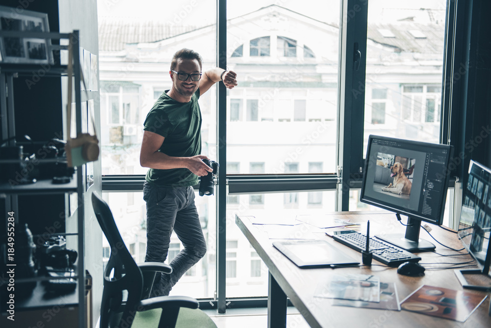 Poster portrait of cheerful unshaven man leaning against window while keeping gadget for creating photos in