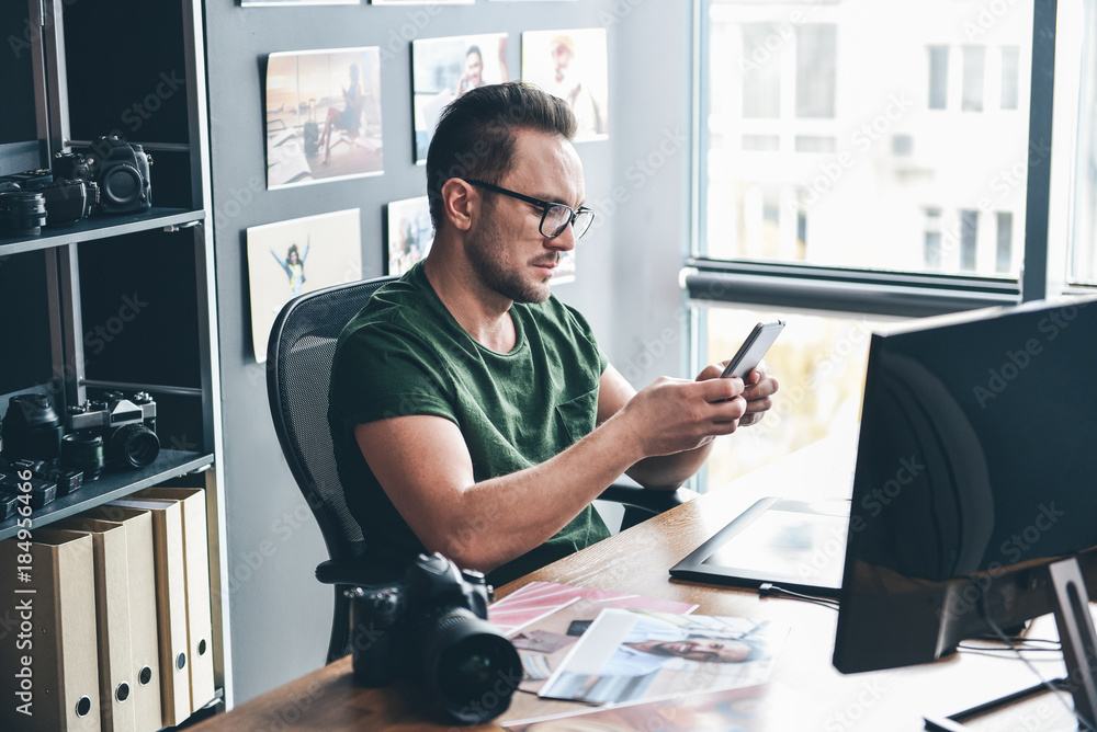 Poster side view serene unshaven male typing message in mobile while sitting at table in modern office. pro