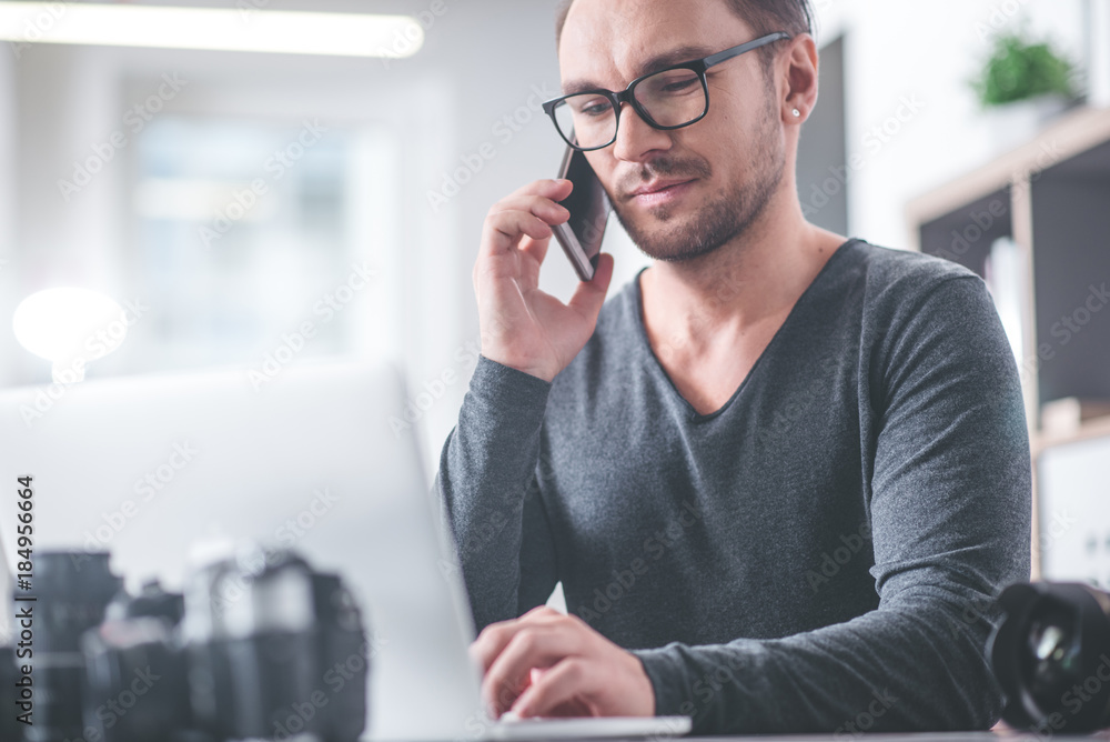 Poster portrait of happy unshaven man chatting by mobile while working with notebook computer in apartment.