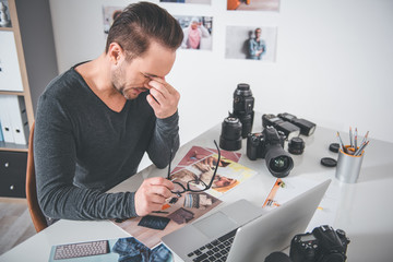 Side view tired bearded man having job with photos and notebook computer at table in office. Occupation concept