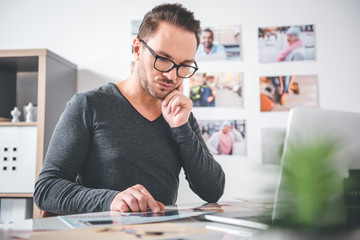 Portrait of serious unshaven male looking at photos while situating at desk in apartment. Job concept