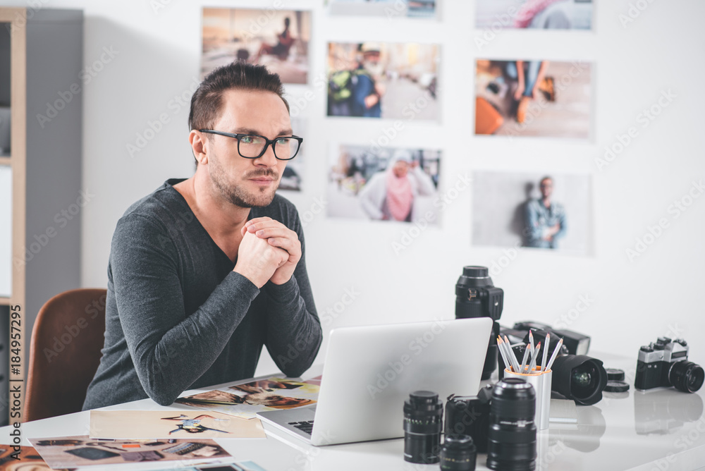 Poster portrait of pensive bearded designer working with digital device and images while sitting in apartme