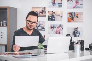Portrait of serene bearded photographer looking at image while sitting at table in modern office. He working at laptop. Job concept