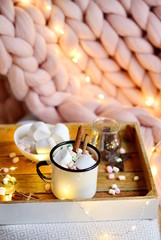 Cup with hot chocolate, bowl with marshmallows, jar with chocolate, pink pastel giant plaid, white chair,  lights, New Year, Christmas concept