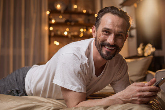 Portrait of cheerful mature man looking at camera with excitement while using mobile phone. He is relaxing in bedroom and smiling