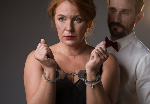 I am your prisoner. Portrait of frustrated middle-aged woman showing her hands locked by handcuffs. Man is standing near her and looking at lady with confidence. Isolated