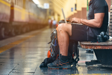 Tourist man with a backpack in the train station working on a laptop. A camera on the bench. Digital nomad traveler concept - obrazy, fototapety, plakaty