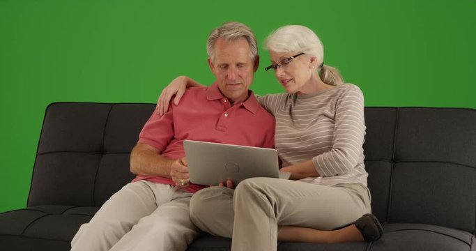Modern senior couple using laptop on couch together on green screen. On green screen to be keyed or composited. 