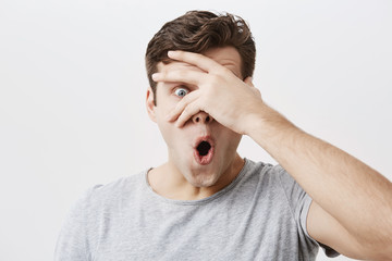 Headshot of goofy surprised bug-eyed young european male student wearing casual grey t-shirt staring at camera with shocked look, expressing astonishment and shock, hiding face behind his palm.