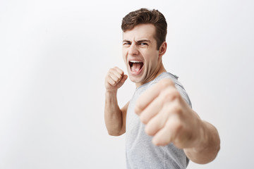 Furious outraged caucasian man shouts in anger, frowns his face, keeps fists, going to defend himself in fight with criminals. Desperate european guy dark hair shows his power, clenches fists.