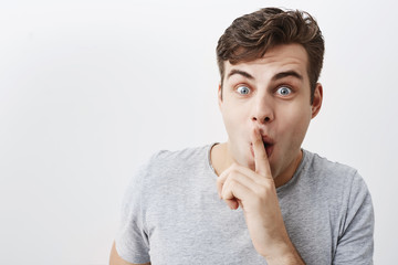 Positive funny man keeps fore finger on mouth, shows silence sign, with popped eyes, asks people to...