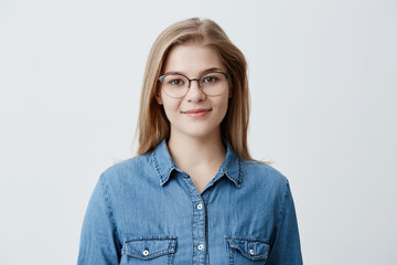 Horizontal portrait of smiling happy young pleasant looking female wears denim shirt and stylish glasses, with straight blonde hair, expresses positiveness, poses against gray background