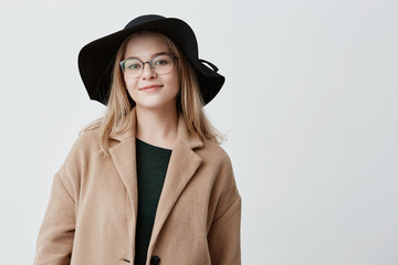 Happy woman with attractive appearance, dressed in trendy coat and black hat, with eyeglasses on, expresses positive emotions, rests after walk in park with friends, has good mood.