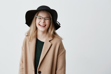 Fototapeta premium Beautiful euphoric teenage girl wearing stylish eyeglasses, black hat and coat, enjoying her life, looking and smiling at camera, full of joy and happiness. People and lifestyle concept