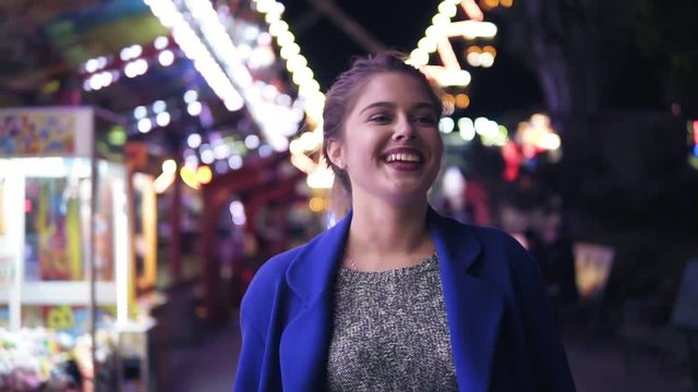Happy young woman walking in amusement park, smiling and laughing. Different attractions on the background. Slowmotion shot