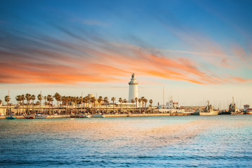Evening panoramic aerial top view to a promenade with palms, shops and yachts and a lighthouse by the sea with dramatic colorful evening sunset sky at the background. Holiday vacation by the sea. - obrazy, fototapety, plakaty