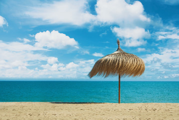 Beach umbrella made of palm leaves standing at the perfect dreamy paradise white yellow sandy bay beach by the blue sea and beautiful dramatic cloudy sky. A place for relax and vacations by the water.