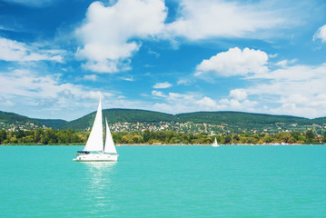 Panoramic view of lake river blue transparent water, a white sport modern luxury yacht sail boat floating and a green shore with forest, hills, villages and beach. Holiday by the water and sail race. - obrazy, fototapety, plakaty