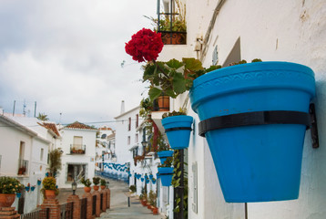 Fototapeta na wymiar Close-up of a blue pot with red geranium at the wall at white narrow street with whitewashed traditional andalusian houses at little touristic town village Mijas, Malaga province, Andalusia, Spain.