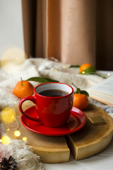 Obraz na płótnie Canvas Close up red coffee cup on wooden cut with winter essentials clementine mandarin, plaid, book and glowing christmas lights on the window sill. Cosiness, holiday morning comfort concept