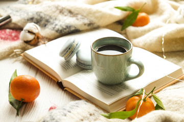 Close up blue coffee cup on the open book with cotton flower, clementine mandarin, french macaroons, plaid, and glowing christmas lights on the window sill. Cosiness, holiday morning comfort concept