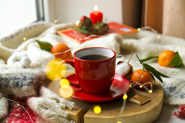 Fototapeta na wymiar Red cup with hot coffee on the wooden cut with clementine mandarin, candle, book and glowing christmas lights on the window sill. Christmas, holiday morning comfort concept