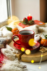 Red cup with hot coffee on the wooden cut with clementine mandarin, candle, book and glowing christmas lights on the window sill. Christmas, holiday morning comfort concept