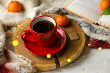 Red cup with hot coffee on the wooden cut with clementine mandarin, book and glowing christmas lights on the window sill. Christmas, holiday morning comfort concept