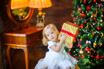 beautiful little girl in a festive dress opens a gift sitting near a Christmas tree.