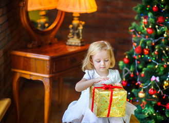Fototapeta na wymiar beautiful little girl in a festive dress opens a gift sitting near a Christmas tree.