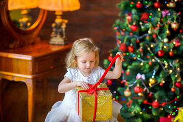 Fototapeta na wymiar beautiful little girl in a festive dress opens a gift sitting near a Christmas tree.