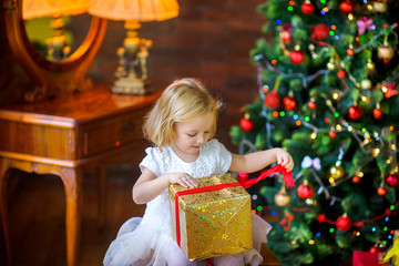 beautiful little girl in a festive dress opens a gift sitting near a Christmas tree.