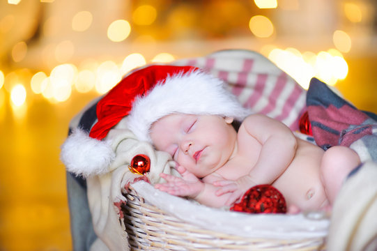 beautiful baby in a Santa Claus hat sipping lying in a basket, against a background of bright festive garlands.