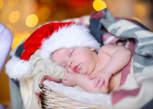 beautiful baby in a Santa Claus hat sipping lying in a basket, against a background of bright festive garlands.
