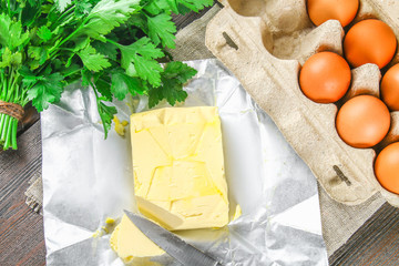 A bar of butter is cut into pieces on a wooden board with a knife, surrounded by milk, eggs and parsley on a brown table. Ingredients for cooking.