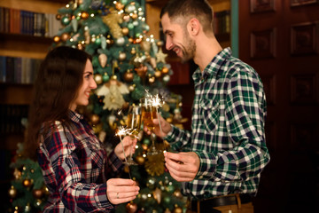 Obraz na płótnie Canvas Merry Christmas and Happy New Year! Attractive young couple is celebrating holiday at home together, drinking champagne and smiling with Bengal lights in hand