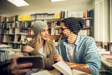 Portrait view of young cheerful happy student love couple with hats sitting at the desk and smiling while taking a selfie in the library.