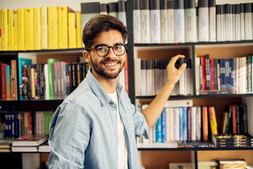 Close up of young cheerful handsome high school student holding a book from bookshelf while looking at the camera in the library.