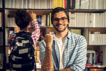 Portrait view of young satisfied smiling bearded hipster student guy in love pointing with a thumb behind him to the curly girl with a backpack in the library.