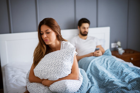 Young beautiful gorgeous sad girl sitting and hugging pillow on the bed in front of her boyfriend with mobile in hands.