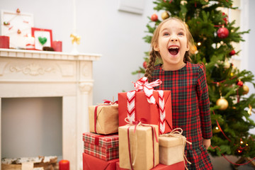 Obraz na płótnie Canvas Portrait of cute little girl with two braids looking away and shouting with delight while standing at pile of Christmas gift boxes, interior of decorated living room with fireplace on background