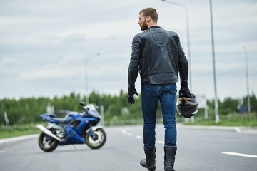 Attractive brutal unshaven young biker man wearing leather jacket boots and gloves sitting...