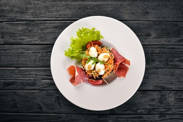 Salad with prosciutto, quail eggs, prunes and fresh vegetables on the plate. On a wooden background. Top view. Free space for text.