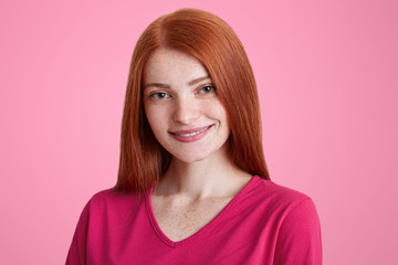 Fototapeta premium Close up portrait of smiling freckled female with straight luxurious red hair, being glad to recieve good job proposal, rejoices her success, isolated over pink background. Facial expressions concept