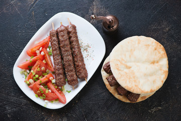 Above view of bbq cevapi or balkan skinless beef sausages served with pitas and tomatoes on a dark brown stone background