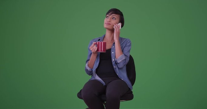 Young african American woman in an office chair drinks coffee and smiles while having a conversation on her smart phone on green screen. On green screen to be keyed or composited. 