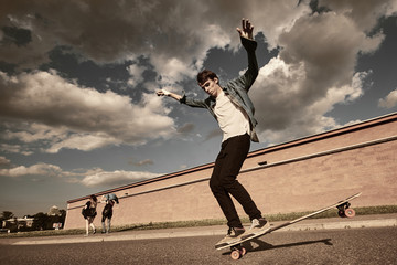 Lifestyle shot of fashionable young male in sneakers, black skinny jeans, white t-shirt and denim shirt enjoying free riding on skateboard in city surroundings against cloudy sky background