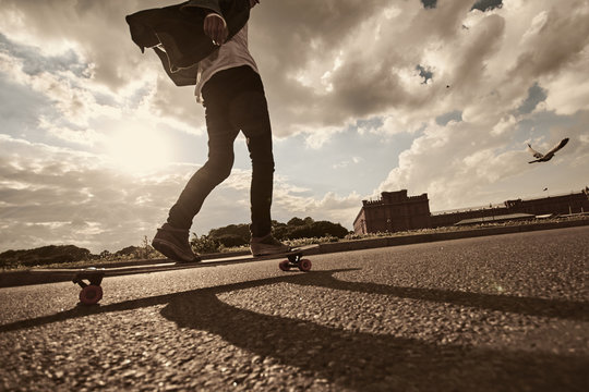 Rear low angle view of unknown skateboarder wearing kicks and skinny jeans sliding on skateboard along paved surface of empty road. Skateboarding, recreational activity and hobby. Flare sun