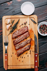 Appetizing roasted meat. Grilled well-done beef steak with with herbs and spices on cutting board and cutlery, flat lay. Restaurant menu concept
