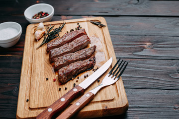 Sliced well-done grilled beef steak on wooden cutting board with rosemary and spices and cutlery, close up. Appetizing meat